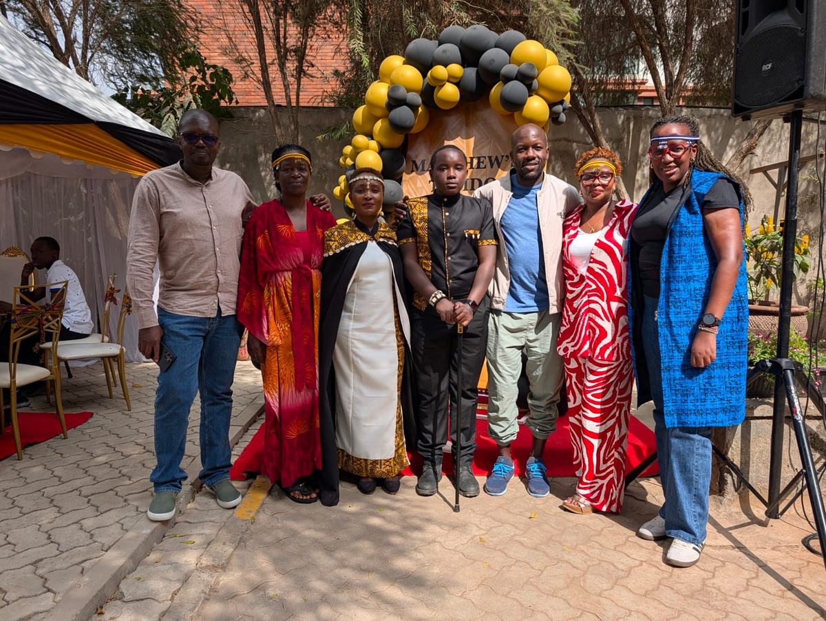 Seven members at an outdoor celebration under a black and gold balloon arch, several wearing beaded headbands.
