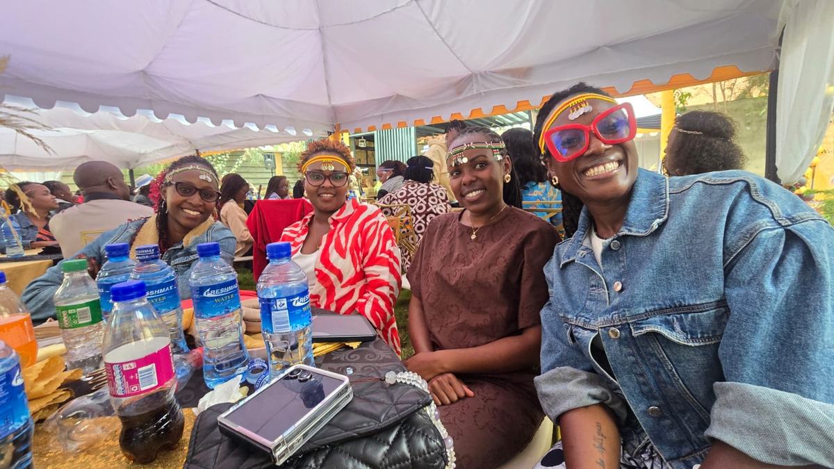 Four members seated together under a marquee in beaded headbands, laughing at the camera.