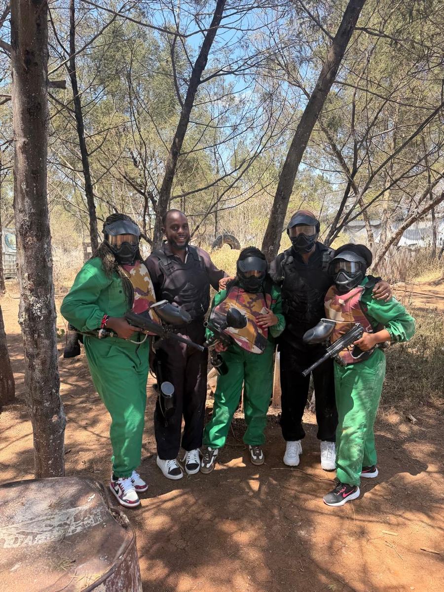 Five members in green paintball overalls and masks among the trees at a paintball range.