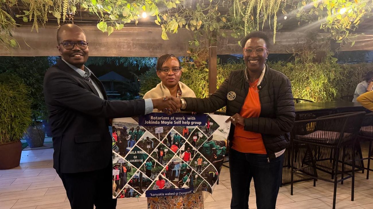 IPP Barrack Odero and PP Jackline Muia shaking hands over a photo banner reading Rotarians at work with Jokinda Moyie Self Help Group, with AG Sylvia Njambi standing between them