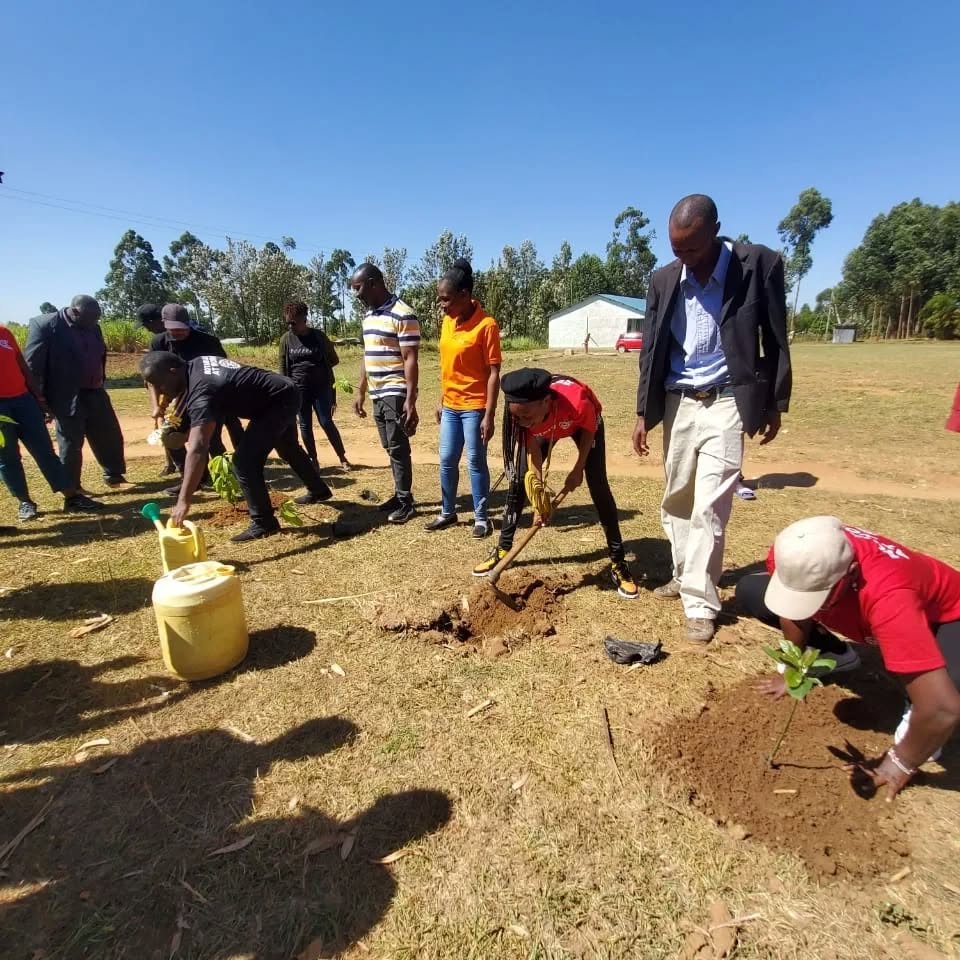 Members and community members planting tree seedlings on open ground, watering cans beside them.