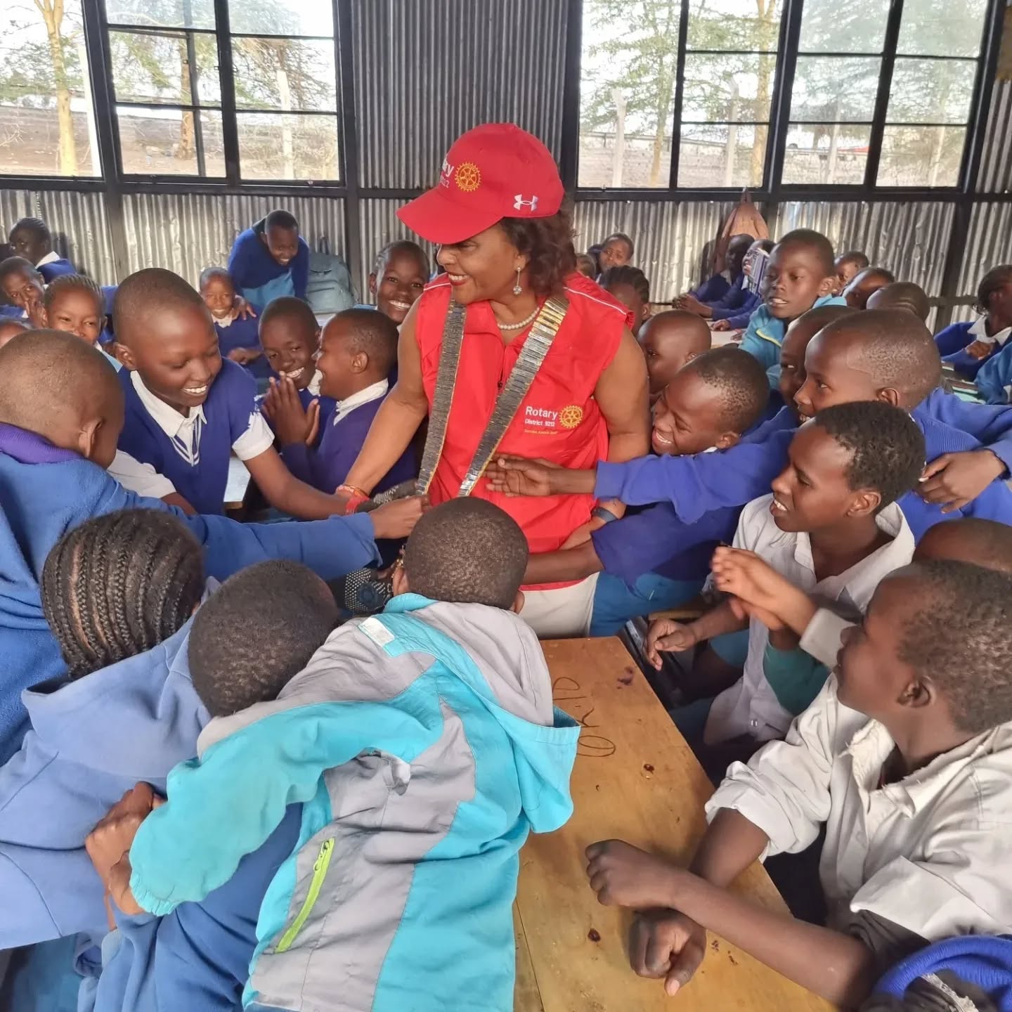 A Rotarian in a red Rotary vest surrounded by primary school pupils in blue uniforms reaching out to greet her.